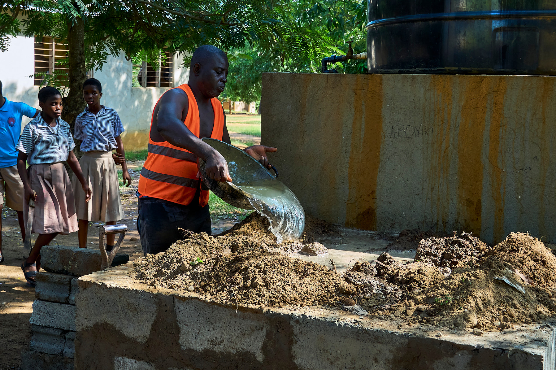 A worker prepares mortar beside a finished drinking water tank at a school, while pupils watch